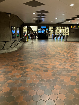 A picture of a metro station pathway. In the background on the left there is a railing protruding from a wall. In the center of the background there is a station manager's kiosk with faregates on both the left and the right side. In the upper right corner of the picture there is a display screen for the time to the next train.